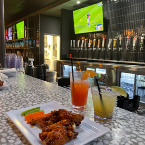 A stack of wings and two cocktails sit on a BowlGames bar counter, framed by a wall of beer taps and TVs broadcasting the game. Come join the party!